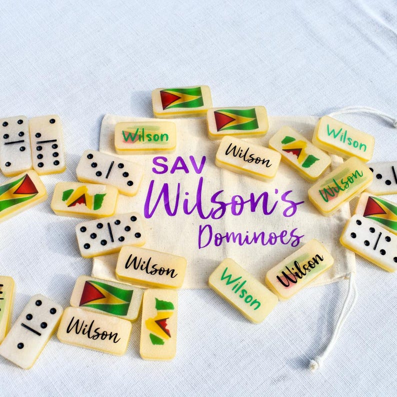 May include: A collection of ivory-colored dominoes with black dots and the word "Wilson" printed on them. Some dominoes feature a flag design with red, yellow, and green colors. A drawstring bag with the text "SAV Wilson's Dominoes" is also present.
