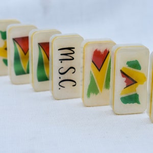 May include: A row of ivory-colored dominoes with a Guyana flag design in red, yellow, green, and black. One domino has the letters "m.s.c." in black. The dominoes are arranged on a white surface.