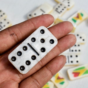 May include: A close-up of a white domino with black dots and a black dividing line, held in a hand. Other dominoes are scattered in the background, some with colorful designs.