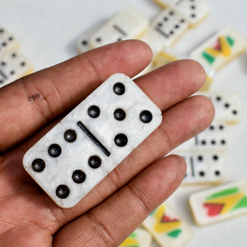 May include: A close-up of a white domino with black dots and a black dividing line, held in a hand. Other dominoes are scattered in the background, some with colorful designs.