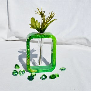 May include: A green glass vase with a clear glass tube inside. The vase is filled with green glitter and has a rectangular shape. The vase is sitting on a white surface with green glass stones scattered around it.