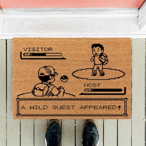 Peut inclure: Un paillasson rectangulaire en fibre de coco beige avec un motif noir pixélisé inspiré d'une interface de combat de jeu vidéo. Le texte indique VISITOR, HOST et A WILD GUEST APPEARED, avec deux personnages et des barres de vie affichés.
