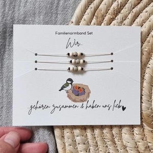 Könnte beinhalten: Ein Set aus drei Armbändern mit Perlen auf einer Karte mit deutschem Text. Die Karte zeigt eine Vogelillustration in einem Nest und die Worte "Familienarmband Set" und "Wir..." in schwarzer Schrift. Die Armbänder haben kleine, helle Perlen.