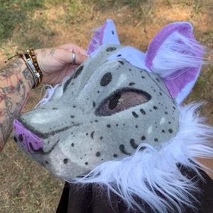Puede incluir: Una máscara de animal de pelo gris y blanco con detalles morados. La máscara tiene una abertura para los ojos de malla y una nariz morada.