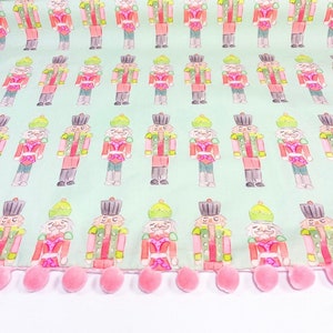 May include: A green fabric with a repeating pattern of watercolor nutcracker soldiers. The fabric has a pink pom-pom trim.