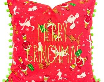 Puede incluir: Cojín decorativo rojo con un diseño temático de Grinch. El cojín presenta el texto "MERRY GRINCHMAS" en verde, junto con personajes de Grinch y copos de nieve. Pompones verdes adornan los bordes, creando un acento festivo.