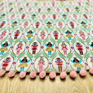 May include: A white fabric Christmas tree skirt with a repeating pattern of nutcracker and fairy figures. The skirt has a pink pom-pom trim.
