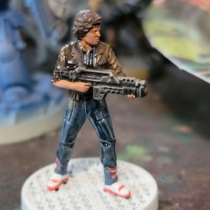 May include: A painted miniature figure of a person wearing a brown jacket, blue jeans, and red shoes, holding a black and silver gun. The figure is standing on a gray and white base.