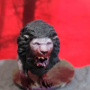 Może przedstawiać: Szczegółowa, ręcznie malowana figurka lwa z ciemną grzywą i zakrwawioną twarzą. Twarz lwa jest w kolorze bladego fioletu, z czerwonymi akcentami wokół ust i oczu. Figurka jest na czerwonym tle.