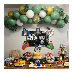 May include: A birthday party table setting with a green, white, and gold balloon arch. The table is decorated with a black and white poster that says "Happy Birthday Giovanni" and features a gorilla. There are also several cupcakes with gorilla toppers, a cake with a gorilla topper, and a variety of snacks and drinks.