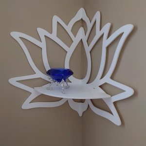 May include: A white, lotus-shaped corner shelf with a small, curved shelf. A large, blue, jewel-like object with silver legs sits on the shelf. The shelf is mounted on a beige wall.