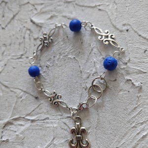Peut inclure: Bracelet argenté orné d'un charm en forme de fleur de lys et de trois perles bleues. Le bracelet comporte des maillons décoratifs et est relié par de petits anneaux. Fond texturé gris clair.