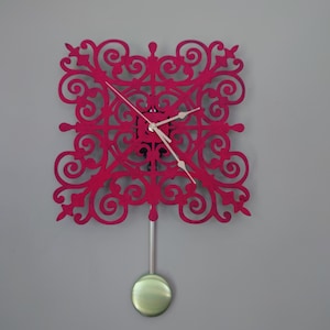 May include: A decorative wall clock with a vibrant magenta filigree design. The clock features a square shape with intricate scrollwork, white clock hands, and a silver pendulum with a green orb. The clock is mounted on a gray wall.