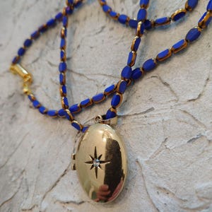 May include: Gold-tone oval locket pendant necklace with a starburst design and a small gemstone. The necklace features blue and gold beaded strands. A classic jewelry piece.