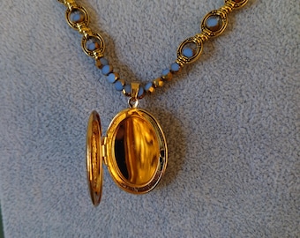 May include: A gold-toned necklace with a pendant locket, open to reveal a golden interior. The necklace features blue and gold beads. The locket is oval-shaped and hangs from a delicate chain, creating an elegant accessory.