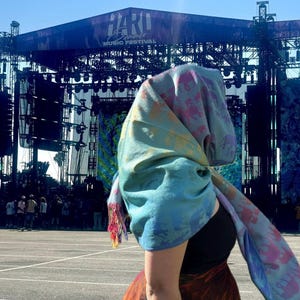 Op de afbeelding: Een persoon draagt een blauw-gele sjaal met olifantenpatronen en een kleurrijke rok. De achtergrond toont een podium met de tekst "HARD Summer Music Festival" onder een heldere blauwe lucht.
