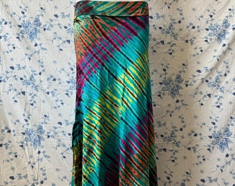 May include: A long, flowing skirt with a vibrant tie-dye pattern. The skirt features diagonal stripes in shades of teal, yellow, pink, and orange. The fabric appears lightweight and has a wide, folded waistband. The skirt is displayed on a mannequin.