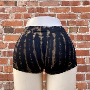 May include: Black and brown tie-dye shorts. The shorts are displayed on a mannequin with a brick wall background. The shorts have a high waist and a short inseam. The tie-dye pattern is vertical and runs the length of the shorts.