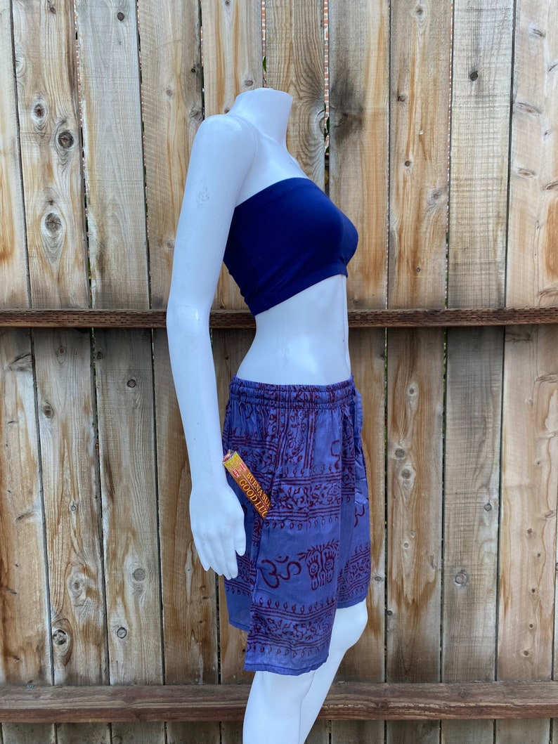 May include: Purple and red patterned shorts with a drawstring waist. The shorts have a pocket on the side with a small orange and yellow package sticking out. The shorts are on a white mannequin against a wooden background.