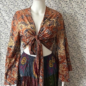 May include: An orange, paisley-patterned tie-front top with long sleeves. The top is paired with a multi-colored, tiered skirt. The fabric has a silky sheen. The outfit is displayed on a mannequin.