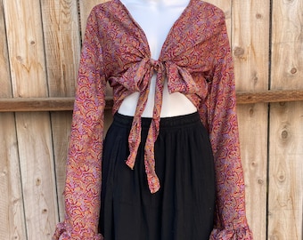 May include: A paisley print tie-front top with long sleeves and ruffled cuffs, paired with a black skirt. The top features a V-neck and a tie closure at the waist. The paisley pattern includes shades of red, purple, and gold.