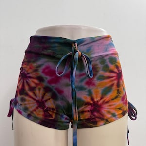 May include: High-waisted shorts featuring a vibrant tie-dye pattern in shades of pink, orange, blue, and dark green. The garment includes a front drawstring closure and adjustable side ties, creating a gathered effect along the outer leg seams.