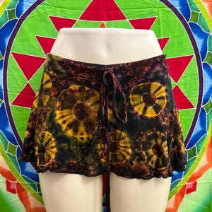 May include: A tie-dye mini skirt in black, brown, and yellow, with a drawstring waist. The skirt has a circular pattern design and a scalloped hemline. The background is a colourful tapestry with geometric shapes.