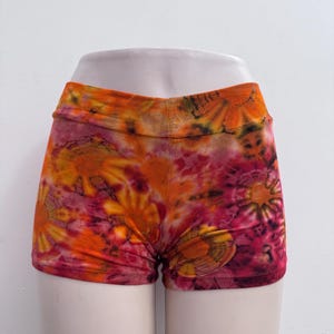 May include: Shorts featuring a vibrant tie-dye pattern in shades of orange, yellow, magenta, and deep red. The garment has a wide, fitted waistband and a short, form-fitting cut that sits at the upper thigh, displaying a circular sunburst design.