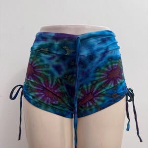 May include: High-waisted shorts featuring a vibrant tie-dye pattern in shades of blue, purple, and dark green. The garment includes adjustable side drawstrings with hanging ties and a matching tie-dye drawstring at the center front waistband.
