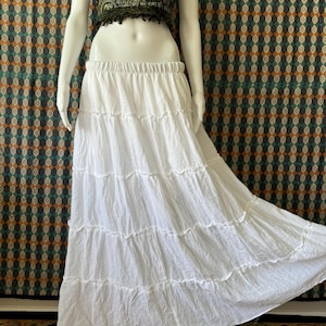 Puede incluir: Una falda maxi blanca de capas con una cintura elástica fruncida. La falda está hecha de una tela ligera y fluida.