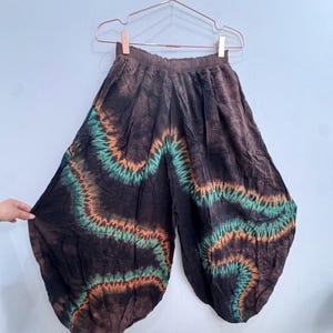 May include: Brown tie-dye trousers with a wide leg design. The trousers feature a wavy pattern in shades of green and orange. The trousers are hanging on a metal hanger against a light blue wall.