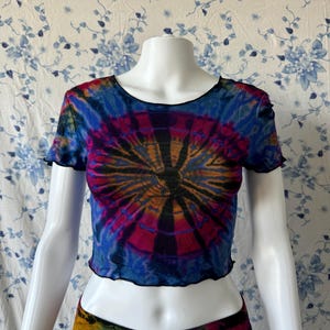 Puede incluir: Un top corto de tie-dye con un patrón circular en tonos de azul, rosa, naranja y negro. El top tiene un dobladillo con volantes y mangas cortas.