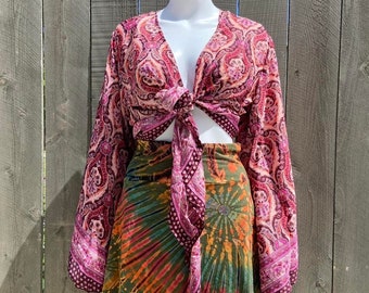 Puede incluir: Una blusa de manga larga con estampado paisley rosa y morado con un nudo en la parte delantera. La blusa se lleva con una minifalda de color verde, naranja y azul tie-dye.