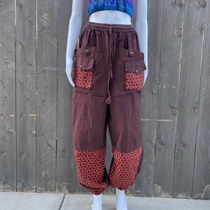 May include: Brown cotton pants with a geometric pattern on the lower legs and pockets. The pants have a drawstring waist and are loose fitting.