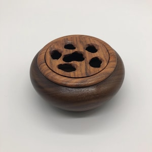 May include: A round wooden bowl with a lid. The lid has several holes cut out of it. The bowl is made of dark wood and the lid is made of lighter wood.