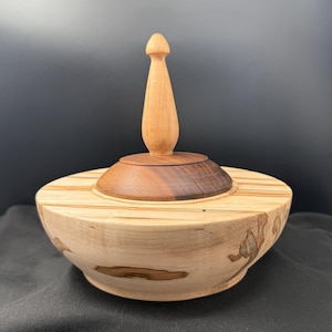 May include: A handcrafted wooden bowl with a lid. The bowl is light-colored with darker wood accents and a decorative finial on top. The lid is a contrasting dark wood. The bowl is likely used for storage or display.