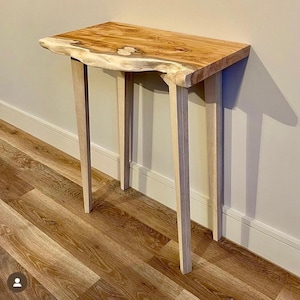 May include: A light wood console table with a live edge top and four slender legs. The table is made of natural wood and has a rustic, organic look.