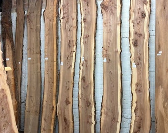 May include: A selection of unfinished, live-edge wooden slabs against a brick wall. The slabs have natural edges, varying in width, with a light brown hue and visible grain. The wood is likely for woodworking projects, with measurements in cm.
