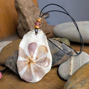 May include: A delicate flower pressed inside a white seashell pendant, with a brown and red beaded cord.