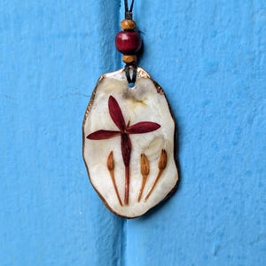May include: A handmade pendant necklace featuring a natural design. The pendant has a white background with a red flower and three brown seed pods. The pendant is edged with gold and hangs from a black cord with a red bead.