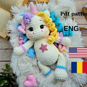 May include: Crocheted unicorn amigurumi toy with a pink heart, yellow and blue yarn, and a star on its chest. The toy is lying on a white fluffy surface. Pdf pattern ENG.