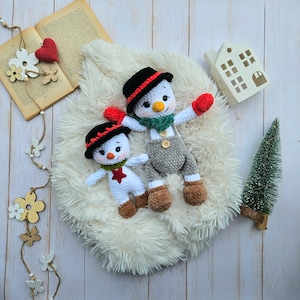 May include: Two handmade crochet snowmen with black hats, red gloves, and brown boots. One snowman has gray overalls. They are lying on a white fluffy surface, with a small Christmas tree and a decorative house in the background. A book and wooden decorations are also present.