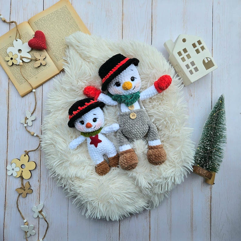 May include: Two handmade crochet snowmen with black hats, red gloves, and brown boots. One snowman has gray overalls. They are lying on a white fluffy surface, with a small Christmas tree and a decorative house in the background. A book and wooden decorations are also present.
