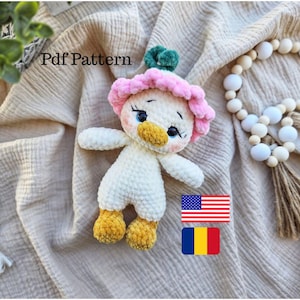 Puede incluir: Una muñeca de pato blanca hecha a mano con ganchillo, con un sombrero de flores rosa y pies amarillos. La muñeca tiene ojos azules y un pico amarillo. El texto "Pdf Pattern" es visible. Las banderas estadounidense y rumana también son visibles.
