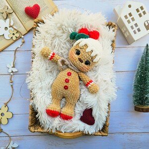 May include: A handmade crochet gingerbread man doll with a white icing head, red and green bow, and red button details. The doll is lying on a fluffy white surface inside a woven basket, with a small Christmas tree and decorative elements.