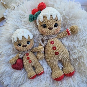 May include: Two crocheted gingerbread man dolls. The larger doll has a white icing-like head, red berry accents, and a scarf. The smaller doll holds a red heart. Both dolls have button details and red accents on their feet and arms. They are resting on a fluffy white surface.