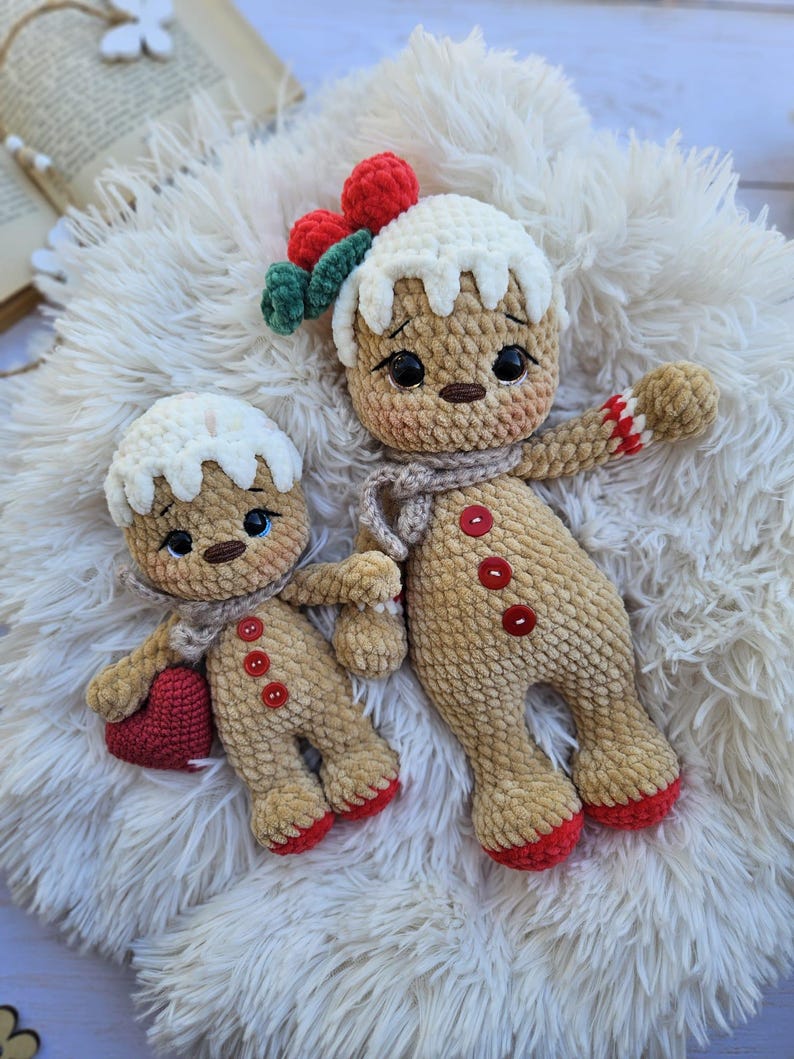 May include: Two crocheted gingerbread man dolls. The larger doll has a white icing-like head, red berry accents, and a scarf. The smaller doll holds a red heart. Both dolls have button details and red accents on their feet and arms. They are resting on a fluffy white surface.