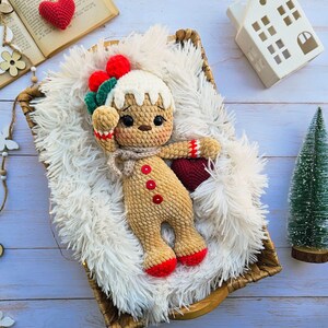 May include: A handmade crochet gingerbread man doll with red and green accents, lying in a woven basket on a fluffy white surface. The doll has a beige body, red buttons, and red feet. A small red heart and a miniature Christmas tree are also in the image.