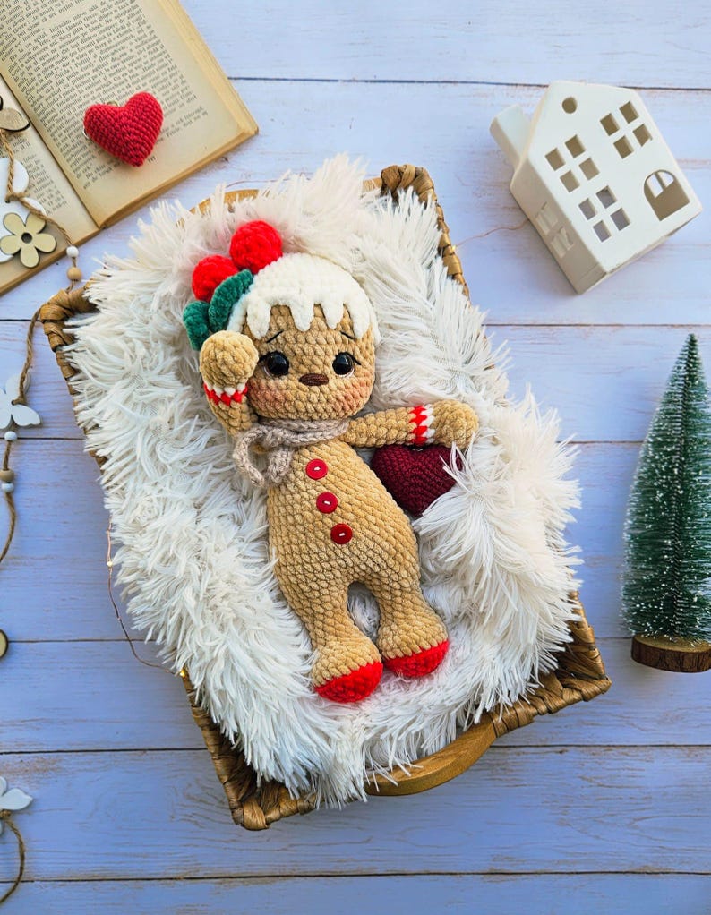 May include: A handmade crochet gingerbread man doll with red and green accents, lying in a woven basket on a fluffy white surface. The doll has a beige body, red buttons, and red feet. A small red heart and a miniature Christmas tree are also in the image.