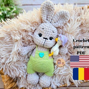 May include: Crochet pattern PDF for a plush bunny toy wearing a green overall and holding a small crocheted duck. The bunny is sitting on a fluffy beige rug. The image also includes the text "Crochet pattern PDF" and flags for the United States and Romania.